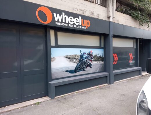 Negozio Wheelup con vetrina che mostra una moto sportiva su strada, ideale per accessori e servizi per motociclisti e viaggi in moto.