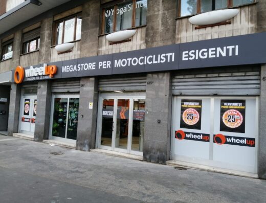 Foto del megastore Wheelup per motociclisti esigenti. L'immagine mostra la facciata del negozio con l'insegna, l'ingresso e promozioni in vetrina. Accessori moto e abbigliamento visibili.