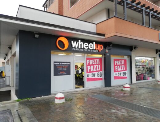 Negozio Wheelup con insegna luminosa e vetrine che mostrano offerte speciali su accessori moto. Orari di apertura visibili, ideale per motociclisti e appassionati delle due ruote.