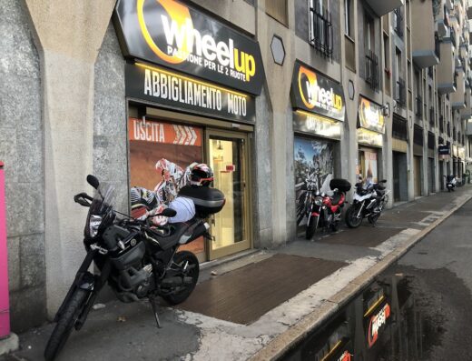 Wheelup: Negozio di abbigliamento moto e accessori a Milano. Moto parcheggiata di fronte all'ingresso, vetrina con esposizione di caschi e abbigliamento tecnico per motociclisti.