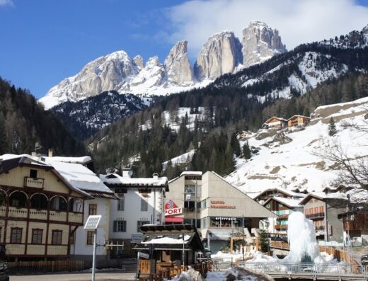 Paesaggio invernale a Campitello di Fassa, Dolomiti: case innevate, montagne maestose, agenzia viaggi Womtravel sullo sfondo.