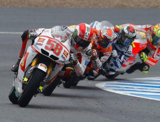 Motociclisti in curva durante una gara di MotoGP, con il pilota numero 58 in primo piano. Caschi colorati e moto da corsa in azione. Esperienza di guida e competizione nel mondo del motociclismo.